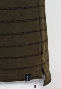 Chemise vert olive à manches courtes avec des rayures horizontales brunes, tissu texturé, fentes latérales et une petite étiquette portant la mention "EST 1993".