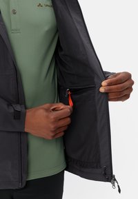 Black jacket with a mesh interior, zippered pocket featuring a red pull tab. Green polo shirt visible underneath. Hands holding the jacket open.