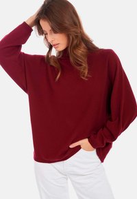Pull oversized bordeaux avec un col montant, manches longues, texture douce, associé à un pantalon blanc. Montre une coupe décontractée et un design drapé.