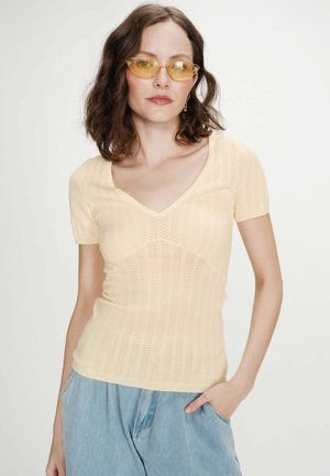 Femme aux cheveux bruns mi-longs portant des lunettes de soleil œil de chat à verres jaunes, un haut en maille jaune pâle à manches courtes et un jean bleu clair.