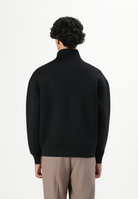 Persona con cabello rizado oscuro de pie de espaldas, vistiendo un suéter negro de cuello alto y pantalones marrón claro, contra un fondo neutro.