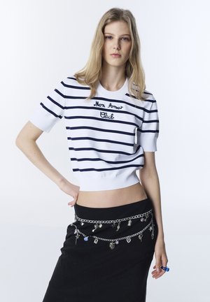 Giovane donna con lunghi capelli biondi che indossa una maglietta bianca a righe blu navy con la scritta "Mon Amour Club" e una gonna nera con catene di charm d'argento.