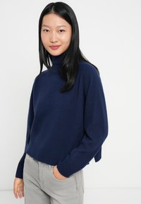Giovane donna con lunghi capelli neri che indossa un maglione a collo alto blu navy e jeans grigi chiari, in piedi con una mano in tasca.
