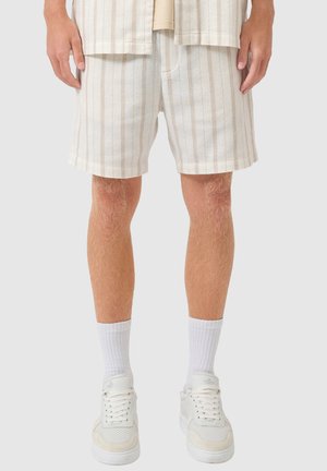 Beżowe spodenki męskie w paski noszone z białymi prążkowanymi skarpetkami i białymi niskimi trampkami, stojące na jednolitym tle.