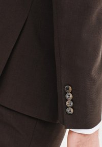 Ciemnobrązowa marynarka o teksturowanym wykończeniu, z czterema guzikami przy mankiecie i widocznym białym mankietem koszuli poniżej.