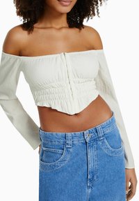 Donna che indossa un crop top bianco a spalla scoperta con maniche lunghe e jeans blu a vita alta, con il torso visibile su uno sfondo bianco.