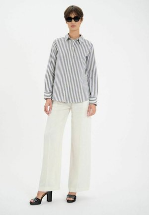Chemise à rayures bleu et blanc ; pantalon large crème ; sandales compensées noires ; silhouette se tenant contre un fond blanc.
