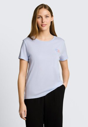 Kobieta z długimi jasnobrązowymi włosami, ubrana w jasnofioletową koszulkę z krótkim rękawem z małym wzorem na piersi i czarne spodnie, stojąca z jedną ręką w kieszeni.