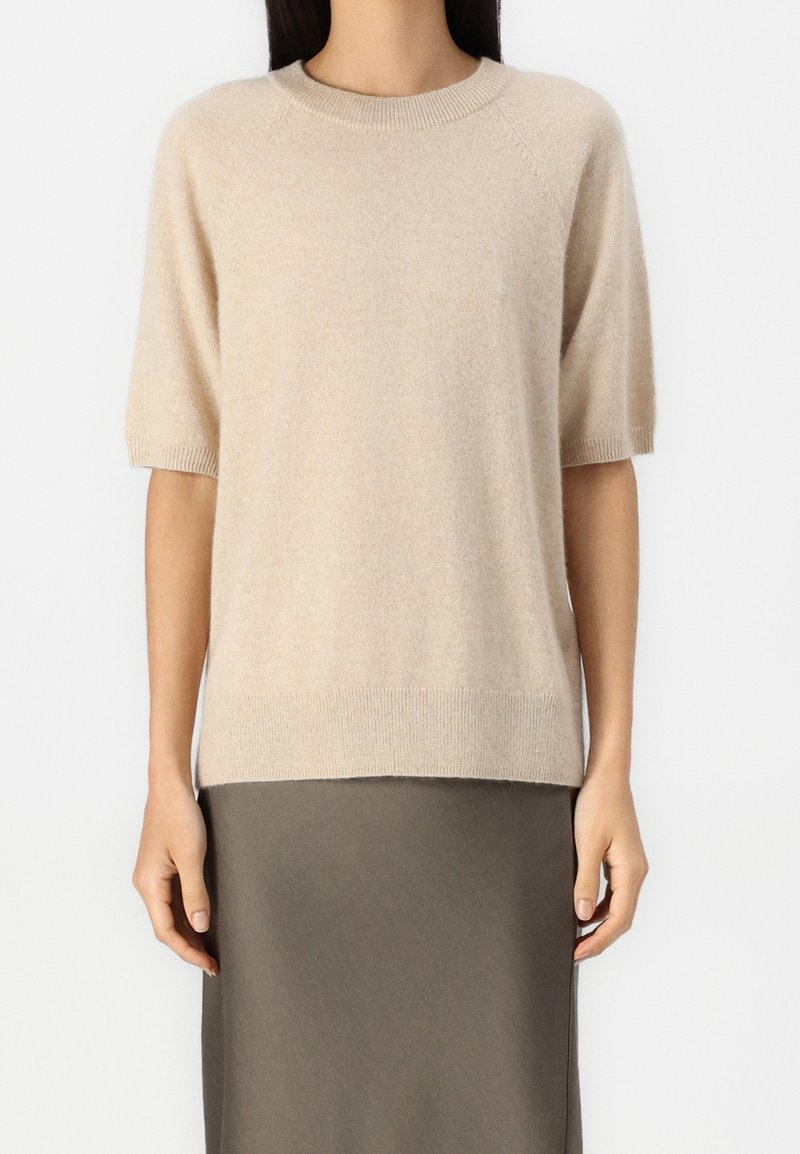 Pull en cachemire beige à manches courtes avec col et ourlet côtelés. Texture douce, coupe décontractée et léger éclat. Associé à une jupe grise.