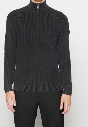 Mężczyzna ubrany w ciemnoszary, prążkowany sweter zapinany na pół zamek błyskawiczny z długimi rękawami i czarne spodnie, stojący na jednolitym tle.