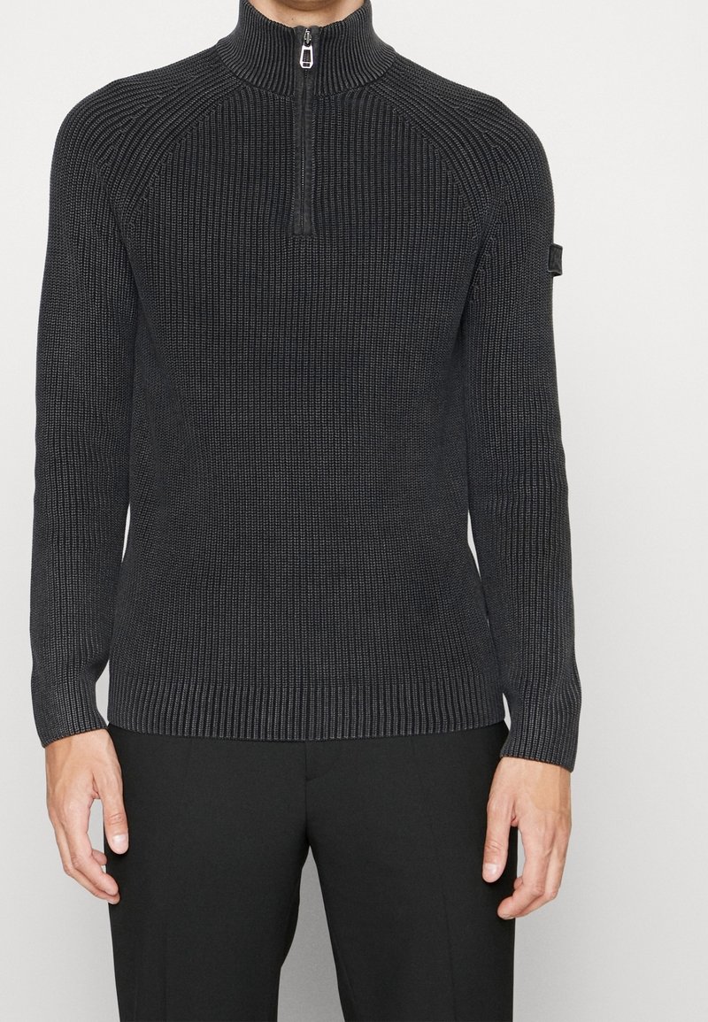 Homme portant un pull gris foncé en maille côtelée à demi-fermeture éclair avec manches longues et un pantalon noir, debout devant un fond uni.