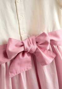 Knöpfe aus hellem cremefarbenem Stoff führen zu einer rosa Schleife an der Taille eines Kleides mit plissiertem Rock, hergestellt aus weicher Baumwolle.