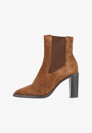 Bottes chevilles en daim marron avec un bout pointu, des panneaux latéraux élastiques et un talon carré texturé. Finition lisse avec des accents de quincaillerie minimalistes.