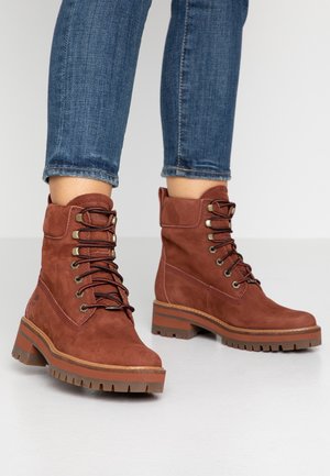 Bottines marron en daim à lacets avec semelles épaisses en caoutchouc, portées avec un jean bleu retroussé sur un sol clair.