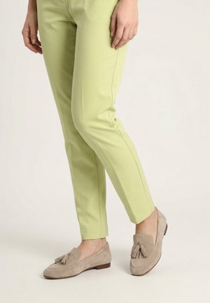 Persoon draagt lichtgroene strakke broek en beige suède loafers met kwastjes, staat met één voet iets naar voren op een witte vloer.