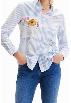 Vrouw draagt een lichtblauw-wit gestreept blouse met knopen, bedrukt met krantenprint en bloemrijke krabbels, gecombineerd met blauwe jeans en handen in de zakken.