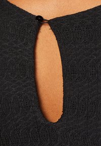 Vêtement noir texturé avec une ouverture en forme de trou de serrure à l'arrière, fermé par un petit bouton noir. Vue rapprochée mettant en valeur un motif complexe.