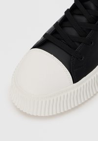 Black sneaker with a white rounded toe cap, smooth leather upper, textured black accents, and ridged white rubber sole. Laces are black.