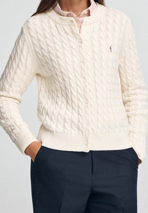 Femme portant un cardigan crème à torsades boutonné sur une chemise rose pâle à col et un pantalon bleu marine foncé, les mains dans les poches.