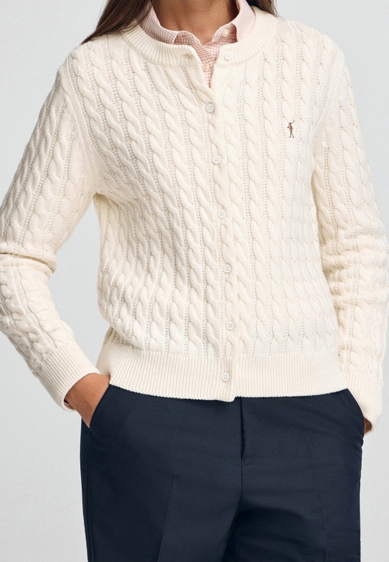 Femme portant un cardigan crème à torsades boutonné sur une chemise rose pâle à col et un pantalon bleu marine foncé, les mains dans les poches.