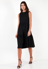 Black, sleeveless dress with a fitted bodice and tiered, flowy skirt. Made from lightweight material. Ankle-strap black heels complete the look.