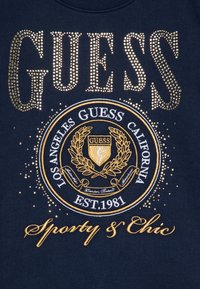 Námornícky sveter s zlatými nitmi, ktoré tvoria slovo "GUESS," obsahujúci vyšívané okrúhle logo, dekoratívne akcenty a slová "Sporty & Chic."