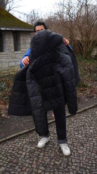 Kék dzsekiben lévő személy nagy fekete hímpuffdzsekit tart egy macskaköves úton kívül, kopár fák és egy közeli kőépület mellett.