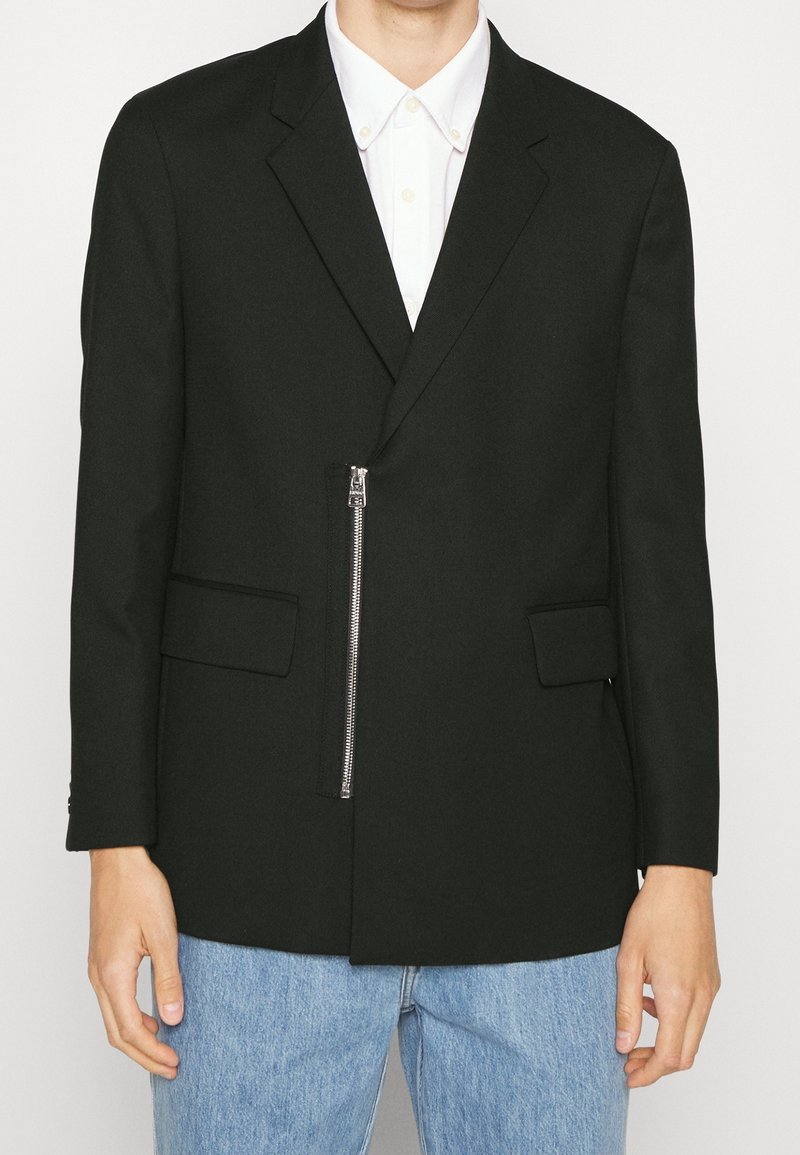 Schwarze Blazer mit einem einzelnen Reißverschluss, zwei Fronttaschen und taillierter Passform. Baumwollmischgewebe mit glatter Textur.