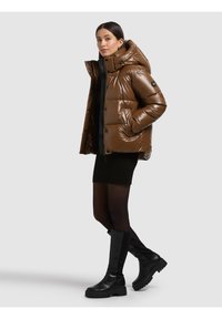 Giacca imbottita marrone con finitura lucida, cappuccio oversize e bottoni a pressione. Indossata sopra un vestito a maglia nero e abbinata a stivali neri al ginocchio.