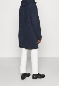 Manteau bleu marine à capuche avec une finition texturée, présentant une coupe droite, un ourlet effiloché, porté avec un pantalon blanc et des chaussures noires.