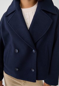 Marinblå, dubbelknäppt ullrock med slagkrage, prydd med två knappar och vertikala ribbade detaljer framtill.