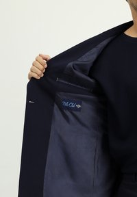 Granatowa marynarka z gładkim, podszytym wnętrzem wyposażonym w kieszeń oraz etykietę z napisem „Polo Club”. Tkanina ma subtelną fakturę.