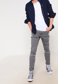 Námořnická košile na knoflíky přes bílý tričko, spárovaná se šedými slim-fit džíny se sešlapanými koleny a ohrnutými manžetami. Bílé tenisky s modrými akcenty.