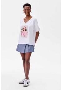 Witte V-hals T-shirt met een roze grafische print op de voorkant, gecombineerd met een blauwe geplooide rok en grijze sneakers. Korte mouwen, ontspannen pasvorm.