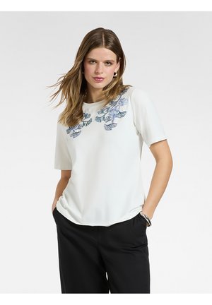 Donna che indossa una maglietta bianca a maniche corte con ricami floreali blu sulle spalle e pantaloni neri a gamba larga, in piedi con le mani nelle tasche.