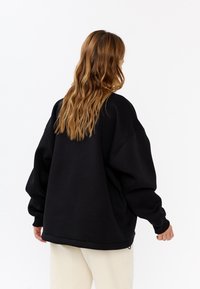 Felpa nera oversize con vestibilità rilassata, spalle cadenti e polsini a coste, realizzata in tessuto morbido. La vista posteriore mostra dettagli minimi.