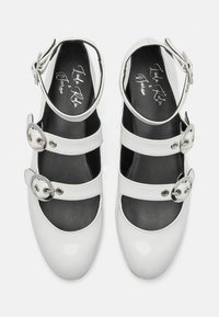 Zapatos Mary Jane brillantes de color blanco con tres correas de hebilla, lados abiertos y plantillas negras. Presentan punta redondeada y correa ajustable en el tobillo.