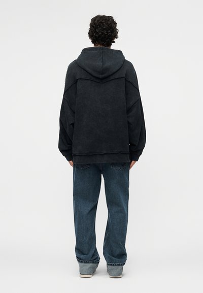 Person mit lockigem Haar, von hinten gesehen, trägt einen dunklen übergroßen Hoodie und lockere blaue Jeans vor einem schlichten Hintergrund.