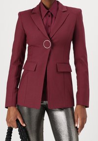 Blazer burdeos a medida con hombros estructurados, detalle de un solo anillo plateado, dos bolsillos delanteros, combinado con una camisa granate y pantalones metálicos.