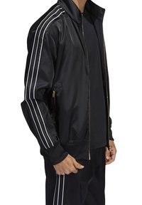 Svart atletisk jacka med vita ränder längs ärmarna, dragkedja och ribbade ärmslut. Tillverkad av släckt, glänsande tyg.