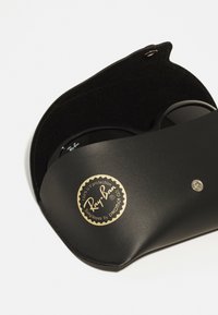 Pătură din piele neagră pentru ochelari de soare, cu o suprafață texturată. Are un logo auriu și un sistem de închidere cu snur argintiu. Ochelarii de soare sunt parțial vizibili în interior.