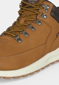 Botte de randonnée couleur sable avec des lacets rayés jaunes et marron, œillets métalliques, col rembourré et semelle en caoutchouc blanche.
