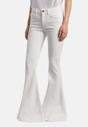 Vrouw die witte, hooggetailleerde, flared jeans draagt met knoop en zakken, gecombineerd met een wit, ingestopt topje, staand tegen een effen achtergrond.