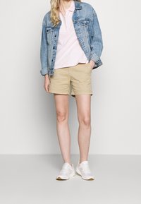 Veste en denim portée sur un polo rose et un short kaki, assortie à des baskets blanches. Les matériaux sont le coton et le denim ; le design est décontracté et ajusté.
