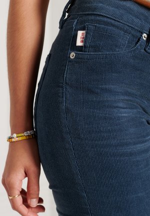 Gros plan d'une personne portant un pantalon en velours côtelé bleu foncé avec une poche avant visible et un bracelet perlé au poignet gauche.