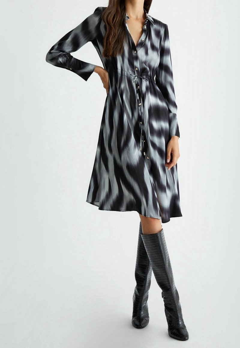 Femme portant une robe boutonnée à manches longues avec un motif noir et blanc, ceinturée à la taille, associée à des bottes noires texturées jusqu’au genou.