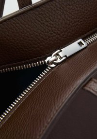 Detail van een bruine leren handtas met een zilveren rits, die een gestructureerd oppervlak en een donkere binnenkant toont. De ritsaanhanger is rechthoekig.