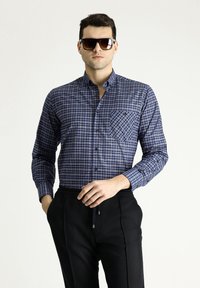 Camicia da uomo con bottoni a quadri blu, dotata di taschino sul petto, maniche lunghe e colletto. Abbinata a pantaloni neri con vita elastica.