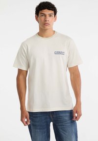 Młody mężczyzna z krótkimi, kręconymi włosami, ubrany w jasnobeżową koszulkę Guess Jeans i niebieskie dżinsy, stojący na jednolitym białym tle.