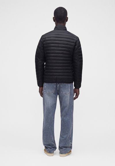 Homme vu de dos portant une veste matelassée noire, un jean ample délavé et des chaussures marron clair sur un fond blanc uni.